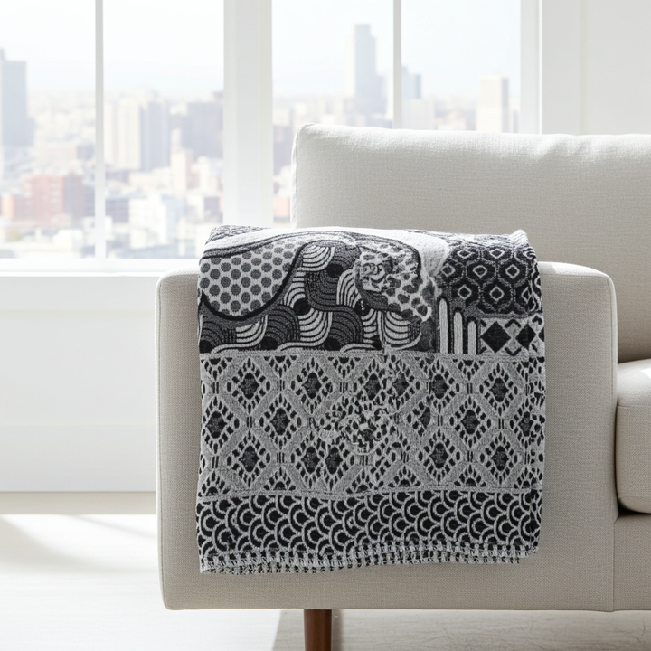A black and white patterned throw blanket with a woven elephant design is draped over the arm of a light beige armchair in a bright, modern room with a city skyline view.