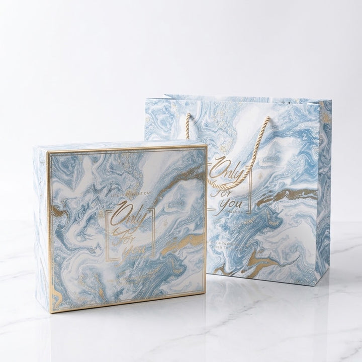A matching gift box and gift bag set with a blue, white, and gold marble pattern, featuring the text &