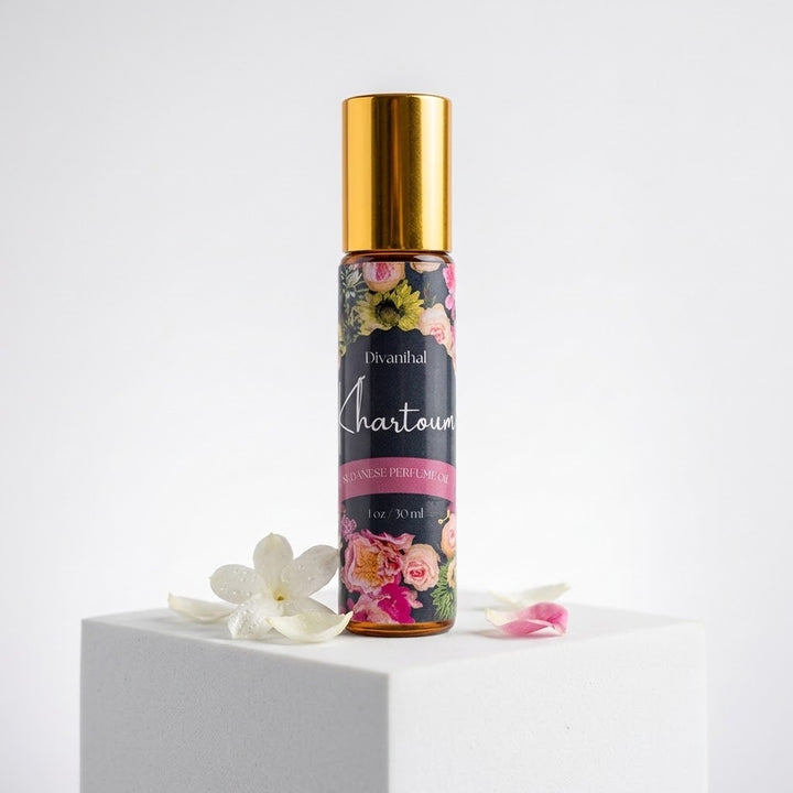 A 10ml roll-on perfume bottle with a shiny gold cap and a floral label, standing on a white pedestal with white flower petals around it against a white background.