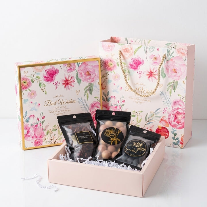 A floral gift set displayed on a white surface, featuring a pink gift box with a gold border, a matching floral gift bag, and three black pouches of bakhoor, cedar balls and incense cones arranged inside the open box with white crinkle paper filler.