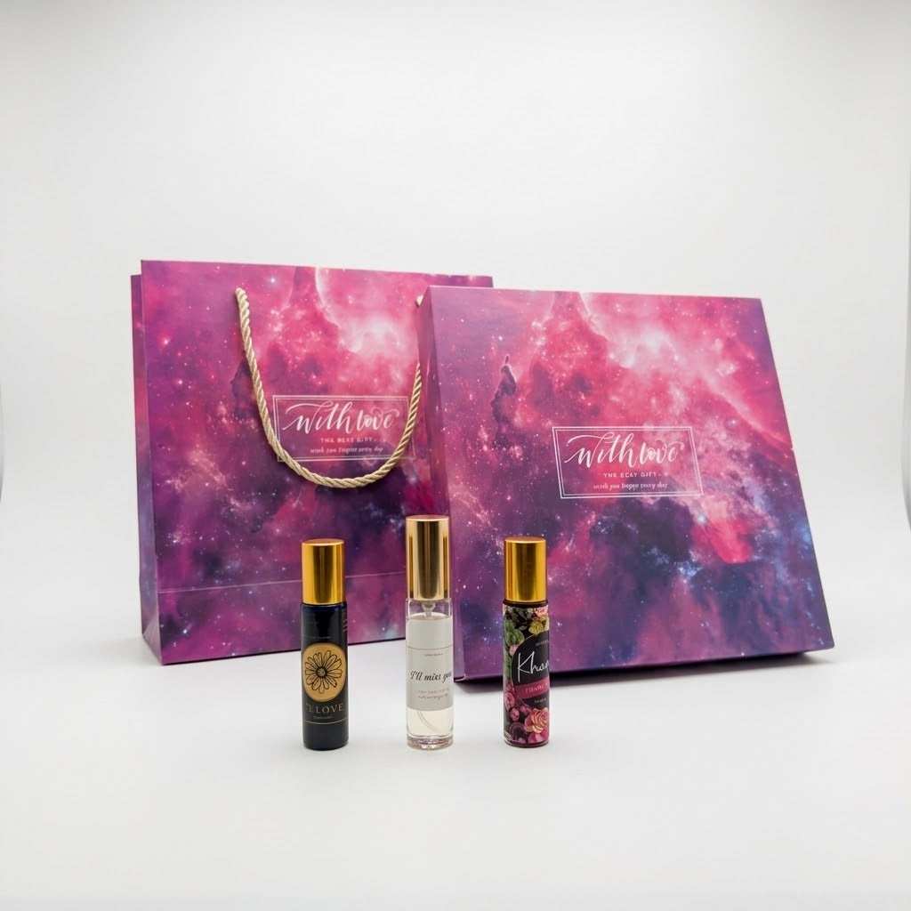 A perfume gift set featuring three small rollerball bottles with gold caps in front of a matching purple and pink galaxy-patterned gift bag and box.