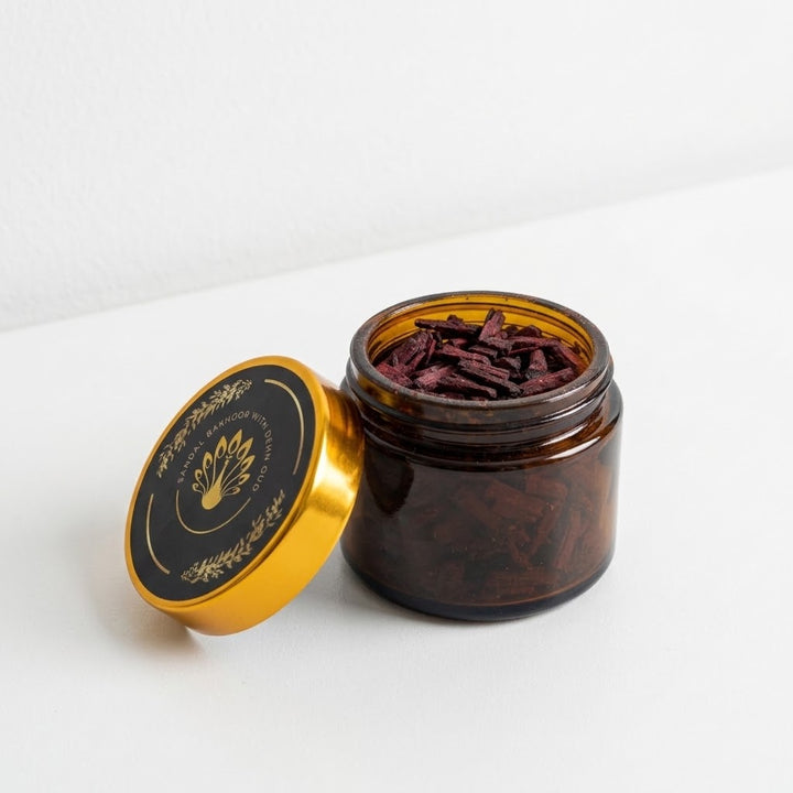 An open amber glass jar filled with dried dark red bakhoor pieces, shown next to its gold and black lid on a clean white surface.
