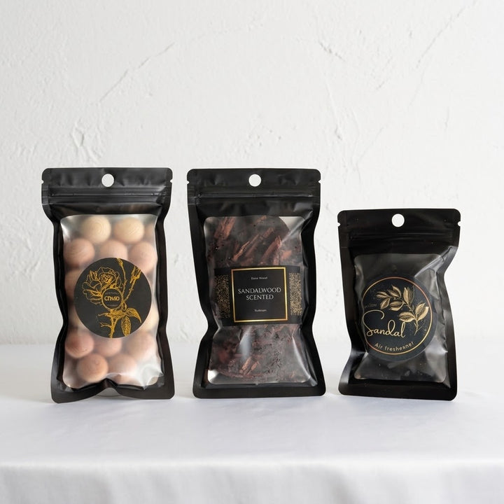 Three black resealable pouches of scented products displayed side by side on a white surface against a white textured wall. The left pouch contains round beige cedar balls with a gold rose label reading &