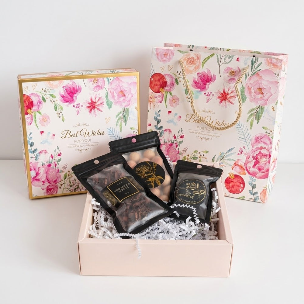 A floral gift set in a pale pink box with white crinkle paper filler, featuring three black pouches of bakhoor, incense cons and cedar balls with gold labels, alongside a matching floral gift box and shopping bag in the background.