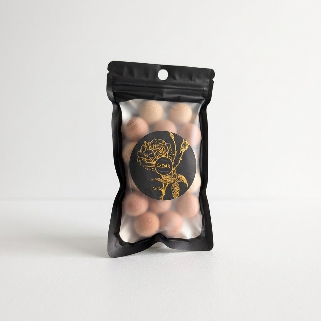 A black resealable stand-up pouch with a clear front window showing round cedar balls inside. A black circular label with gold floral artwork and the word &