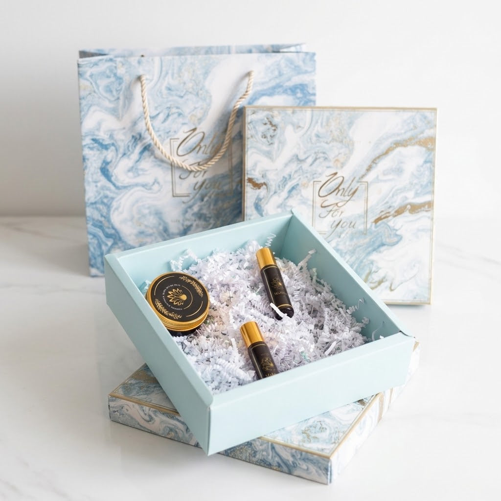 A gift set presentation with a light blue open box filled with white crinkle paper, containing two small dark bottles with gold caps and a round gold-labeled tin. Behind it are matching blue-and-white marbled gift bag and box packaging on a clean white surface.
