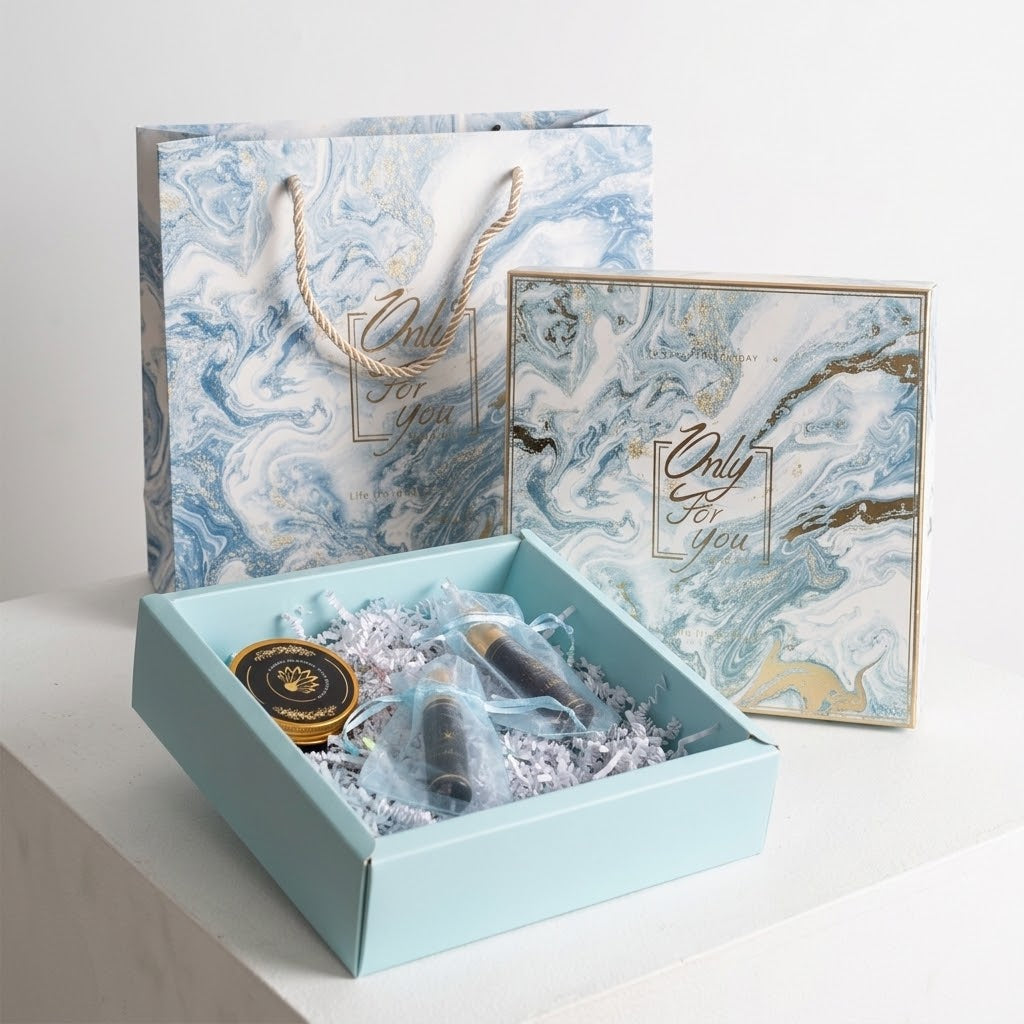 A gift set displayed on a white surface, featuring a light blue box filled with silver crinkle paper and a small black-and-gold item, alongside a matching blue-and-white marbled gift bag and square box with gold foil script reading 'Only For You'.