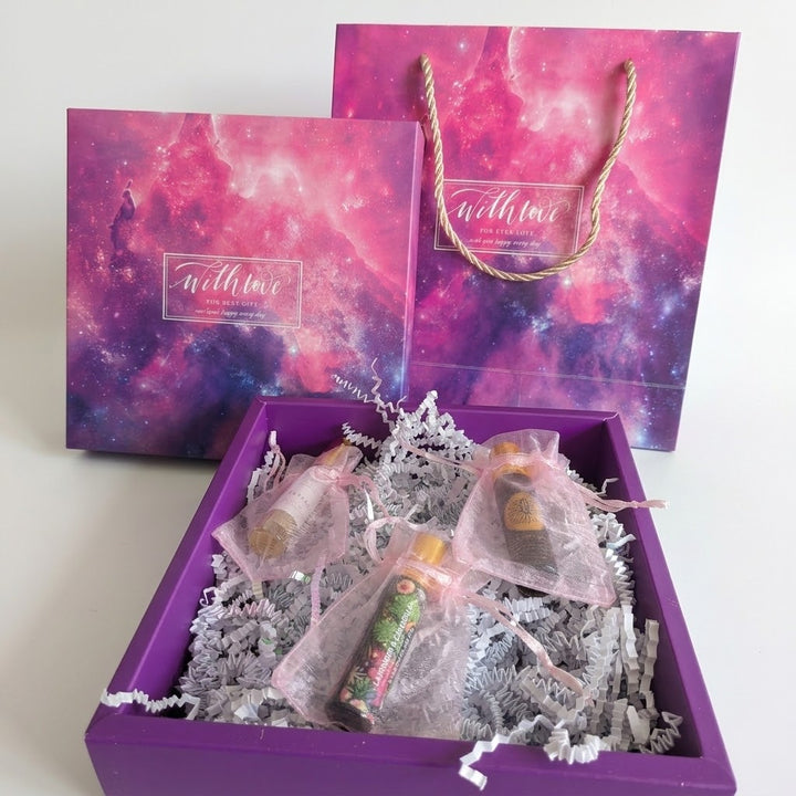 A purple gift box filled with small wrapped bottles and tissue paper filler, shown with matching branded gift bag and box lid in front of it. The packaging has a pink and purple galaxy print, and the front bag has twisted rope handles.