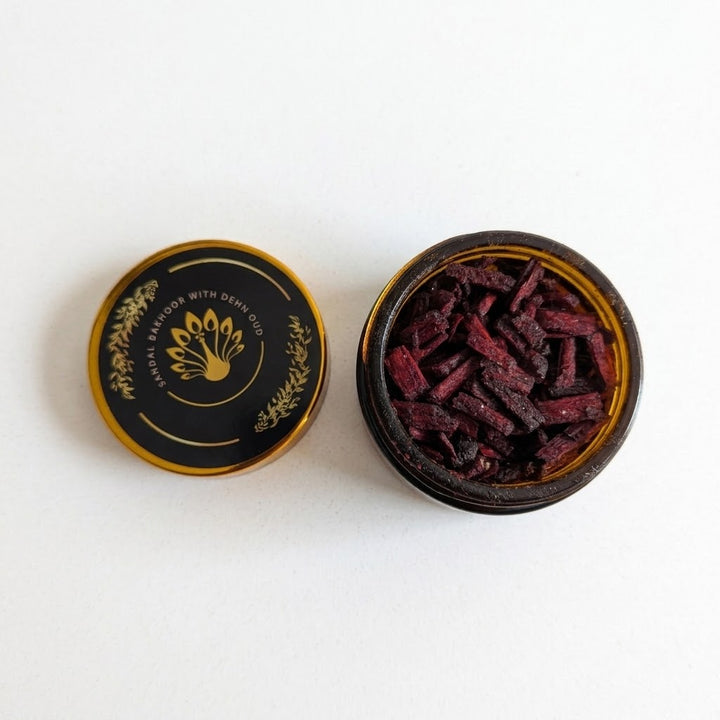 An open round amber glass jar filled with dark reddish-brown dried bakhoor pieces, shown next to its black lid with gold lettering and a lotus flower emblem on a white background