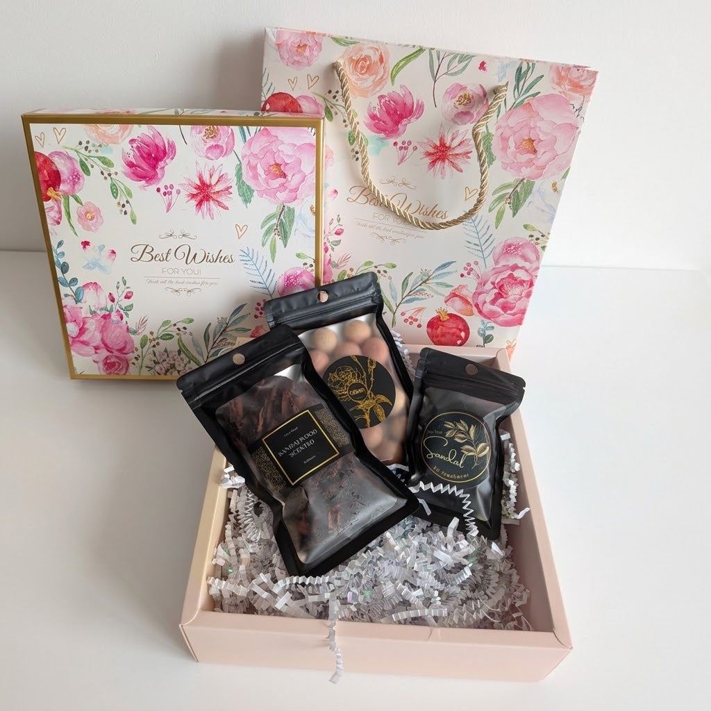 A floral gift box set on a white surface, featuring a pink and gold gift box with shredded paper filler, three black pouches filled with incense cones, bakhoor and cedar balls with gold labels, and a matching floral gift bag in the background.
