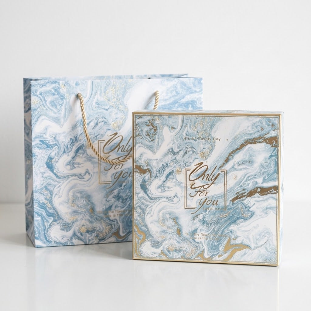 A square gift box and matching paper shopping bag with a blue, white, and gold marble pattern, featuring the text &