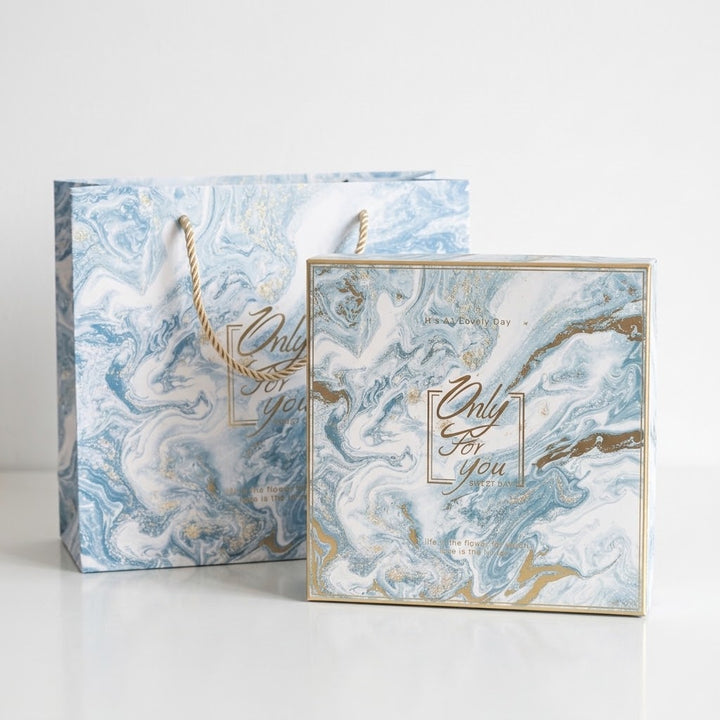 A square gift box and matching paper shopping bag with a blue, white, and gold marble pattern, featuring the text &