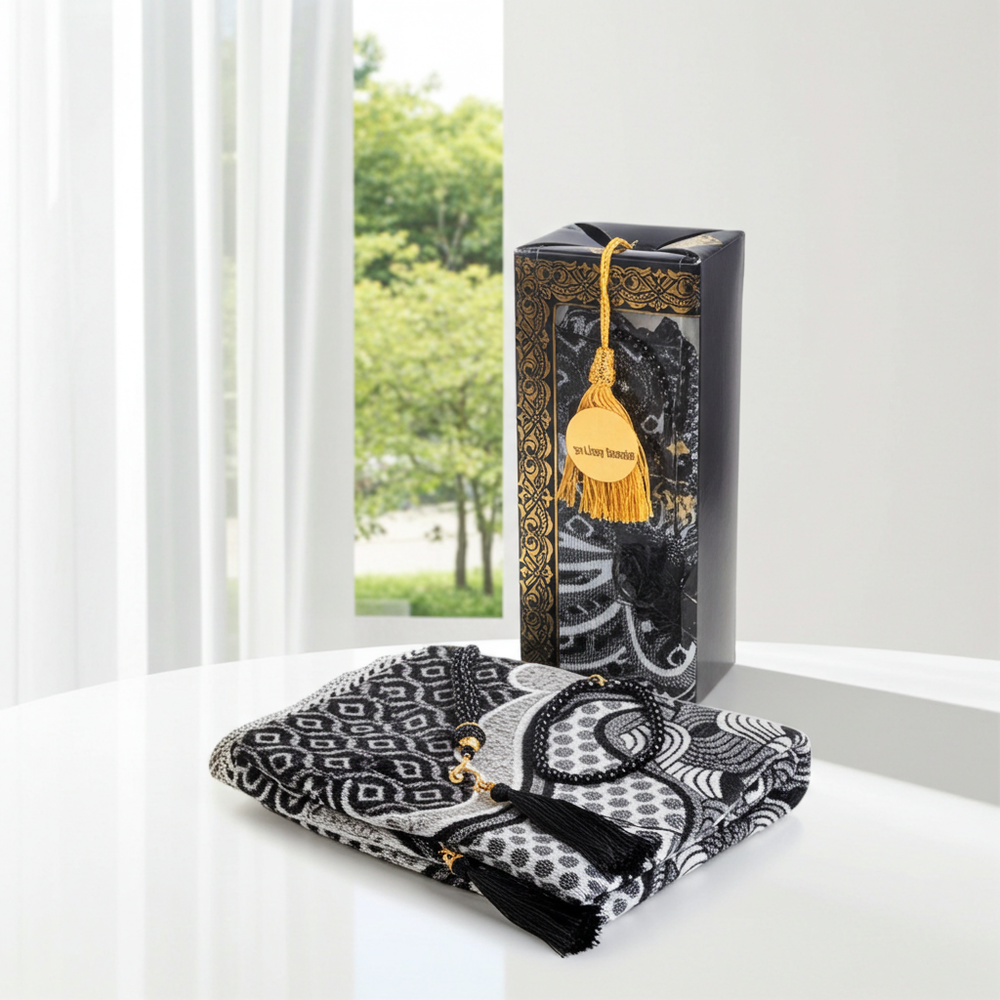 A folded black and white patterned scarf with a gold tassel and beaded cord rests on a white table in front of its black gift box, which features a gold tassel tag reading 'Anjing Jannah'.