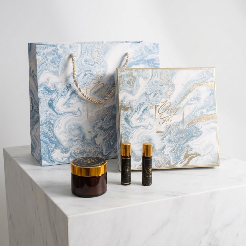 A gift set featuring a blue and white marble-patterned shopping bag, a matching square gift box with gold foil accents, a dark amber jar with a gold lid, and two small black perfume or cosmetic bottles with gold caps, arranged on a white marble pedestal against a light gray background.