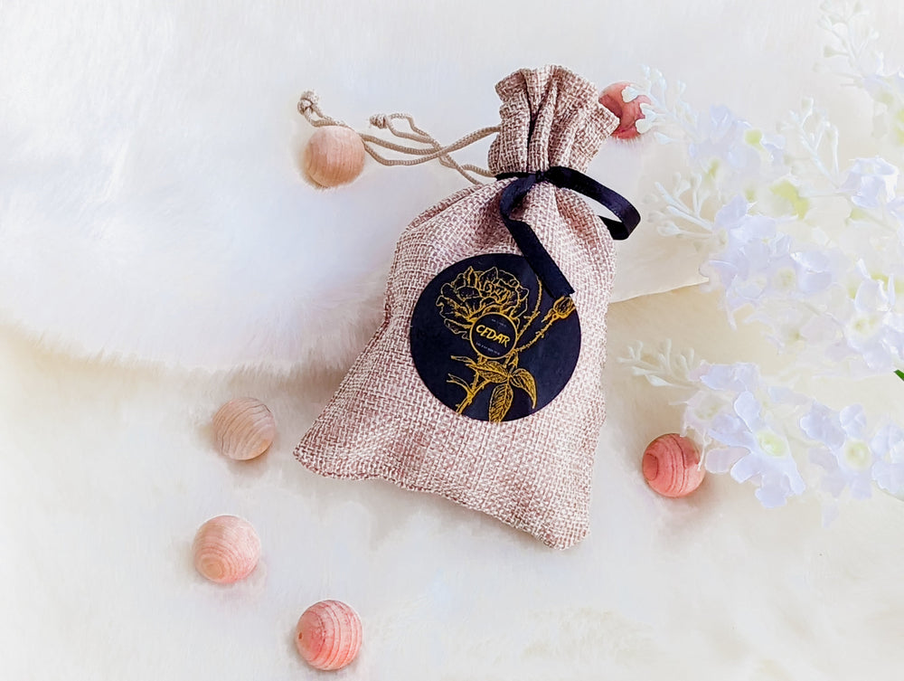 A small burlap sack with a drawstring closure and a black label featuring a gold floral design, set against a pink background.